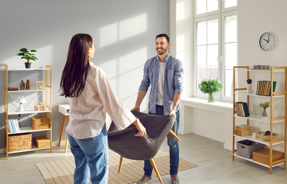 Young,Couple,Carrying,Armchair,Into,Their,Living,Room.,Smiling,Man